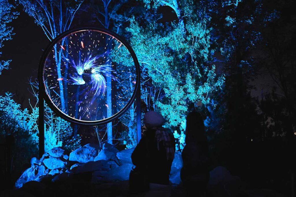 A person stands before a blue-lit sculpture, highlighting the artwork's intricate design and vibrant illumination.